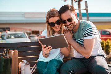 Wall Mural - Young smiled couple is sitting on bench outside, wearing glasses while they enjoy sunny weather