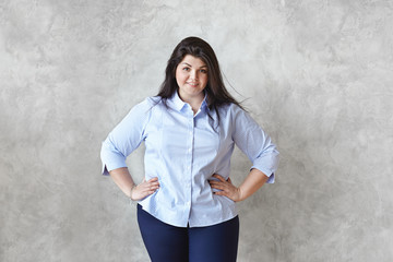 Portrait of attractive friendly looking young big-boned overweight brunette woman model wearing blue striped shirt and skinny jeans posing at blank wall, keeping hands on her waist and smiling