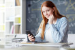 © pressmaster - Smiling student looking at camera while sitting by her desk with smartphone and messaging at break