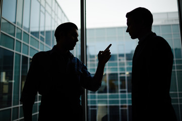 Silhouettes of two young men discussing working points by large window of modern business center