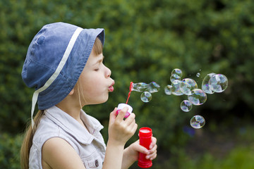 Profile of pretty small preschool blond girl with funny serious expression blowing colorful transparent soap bubbles outdoors on blurred green summer background. Joy of careless childhood concept.