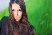 Portrait Of Beautiful Blue-eyed Girl Brunette With Long Hair, Disheveled By Wind, On Blurred Background Of Green Grass Looking At The Camera, Closeup.