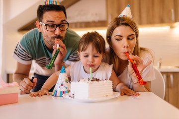 Wall Mural - Cute little girl is having little birthday party with her parents and she is blowing candle on a birthday cake