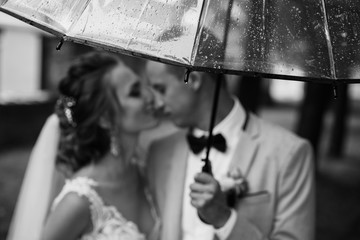 Wedding rain. Bride and groom in the rainy weather are covered with a transparent umbrella and kisses. Selective focus on rain drops in umbrella