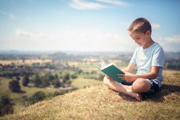 Boy sitting on a hill reading a book in a meadow