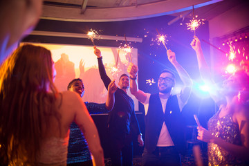 Wall Mural - Group of trendy young people celebrating holiday in nightclub burning flaming sparklers on dance floor, copy space