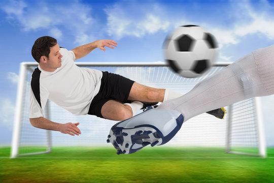 Composite image of football players tackling for the ball against football pitch and goal under blue sky