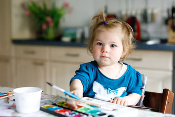 Wall Mural - Cute adorable baby girl learning painting with water colors. Little toddler child drawing at home, using colorful brushes