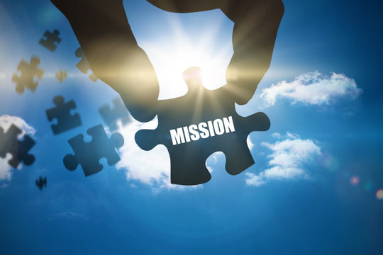 The word mission and hand holding jigsaw piece against bright blue sky with clouds