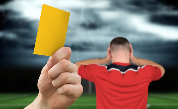 Hand holding up yellow card against football pitch under stormy sky and player