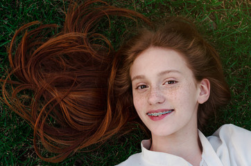 a red-haired girl with beautiful brown eyes and braces on her teeth lies on the green grass and smil