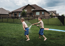 Side View Of Shirtless Boy Spraying Water On Brother With Garden Hose At Park