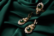 Beautiful Golden Ring And Pair Of Earrings With Green Emerald And Diamonds Gemstones On A Green Satin Background. Luxury Female Jewelry, Close-up. Selective Focus