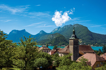 Wall Mural - View of houses and belfry with blue sky mountains landscape on background, in the village of Talloires. A lovely village next to the Lake of Annecy. Department of Haute-Savoie, southeastern France.