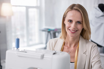 Wall Mural - Customer oriented focus. Positive female tailor grinning to camera while using sewing machine