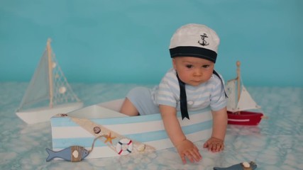 Sticker - Cute toddler baby boy in wooden boat, playing with fishes, starfish and binoculars as sailman, studio indoors shot