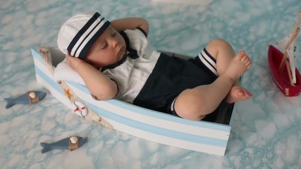 Wall Mural - Cute toddler baby boy sleeping in wooden boat with fishes, starfish and binoculars, dressed as sailman, indoors shot