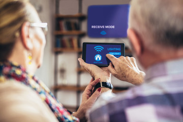 Elderly couple sitting comfortably on a sofa with their backs holding remote home control system on a digital tablet