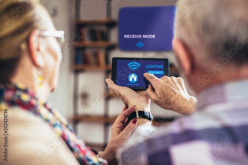 Fotografía Elderly couple sitting comfortably on a sofa with their backs holding remote hom