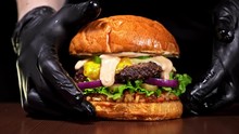 Burger is cooking on black background in black food gloves. Very luscious air bun and marbled beef. Restaurant where each burgers is cooked by hand. Not made ideal. Not conveyor. Looks real, loving