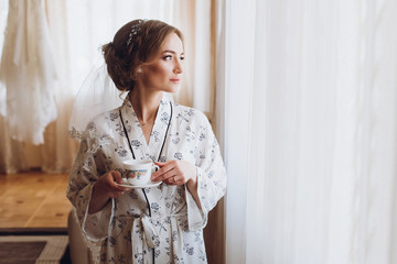 Gorgeous bride in elegant robe drinking coffee in luxury hotel room near window, morning before the wedding preparation