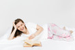 © ViDi Studio - Young happy woman lying in bed with white sheet, pillow, blanket on white background. Beauty female spending time in room, reading book, enjoying of rest. Relax, good mood concept. Place for text.