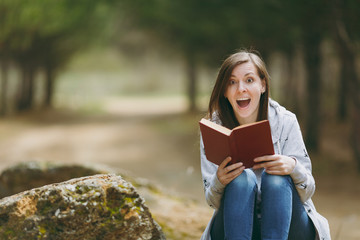 Young overjoyed shocked beautiful woman in casual clothes sitting on stone studying reading book in city park or forest on green background. Student learning, education. Lifestyle, leisure concept.