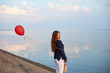 Portrait of young woman with red air balloon and present bag near the calm sea or lake shore. Clouds are reflected on the smooth water surface. Girl on her birthday. Copyspace. Holiday concept.