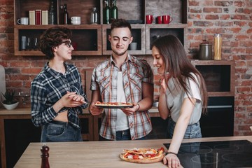 Wall Mural - Confident young man with pizza and delighted girls praise their friend for cooking. Success, achievement, friendship, togetherness, cooking, people and food concept