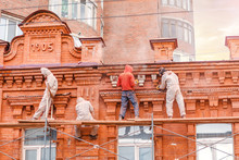 Unrecognizable Workers In Protective Clothing Are Engaged In The Restoration Of The Old Brick Building, Concept Of Hard Severe And Harmful Working Conditions