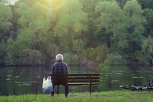 Old Man Sitting On Bench Near River And Watching Sunset. Senior Man Sitting Alone In Park.