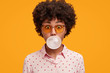 Playful funny African American woman blows chewing gum, spends spare time with friends, wears shirt and sunglasses, isolated over yellow background. Shocked female inflates bubble gum in studio