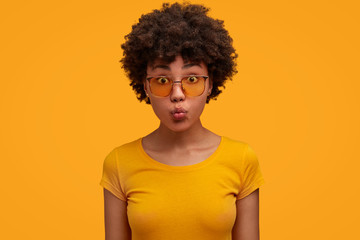 Isolated shot of pretty dark skinned female student makes grimace, keeps lips round, wears casual t shirt and shades, isolated over yellow background. Curly African American woman enjoys summer rest