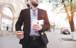 © bodnarphoto - A businessman walking down the street with a snack and coffee in his hands. A busy man eats a sandwich on the road to work. Breakfast. Business concept.