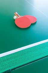 View of two red table tennis bats or rackets with a white ball on a green table with a net