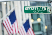 Close-up View Of Green Street Sign Depicting It Is Rockefeller Plaza In Midtown Manhattan, New-York. Blurred American Flags In The Background