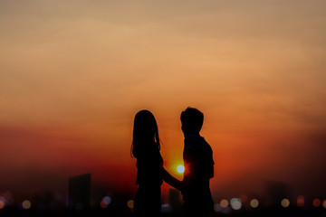 two people silhouetted against a bright orange sunset. They are holding hands, with a city skyline in the background.