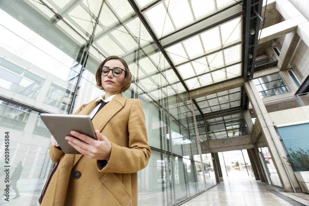 Donna d'affari con occhiali da vista vestita con cappotto beige utilizza il suo tablet in un contesto esterno aziendale 