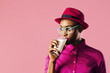© Carlos David - Portrait of a young woman with glasses holding and drinking a glass of milk, looking to the side,, isolated on pink studio background