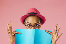 The Reader - Portrait Of A Young Woman With Glasses Holding A Book And Peeking Up From Behind It With Joy, Isolated On Pink Studio Background 
