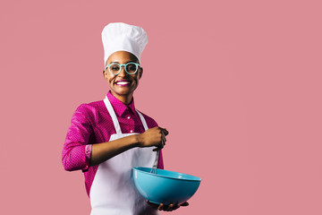 Portrait of a very happy and smiling young woman in cooking hat and apron mixing a bowl with a whisker, isolated on pink studio background 