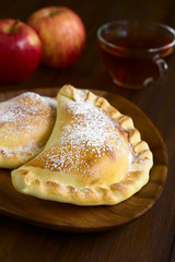 Chilean apple empanada with icing sugar, cup of tea in the back, photographed on dark wood with natural light (Selective Focus, Focus one third into the image)