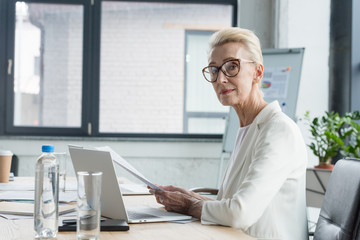 Wall Mural - attractive senior businesswoman in eyeglasses holding documents and looking at camera in office