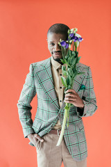 Wall Mural - smiling african american man in jacket holding bouquet of flowers, isolated on red