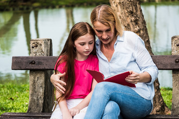 Wall Mural - Mother and daughter reading a book together in a park. Family, lifestyle and education concept