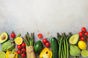 Different organic vegetables - asparagus, tomatoes cherry, avocado, artichoke, pepper, lime, lemon, salt on gray background. Food fame with copy space, top view. Raw, vegan, vegetarian concept.