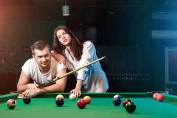 A guy and a beautiful girl are playing billiards, a guy is teaching a girl to play billiards. Dark background. Pleasant pastime, family rest, entertainment, holiday.