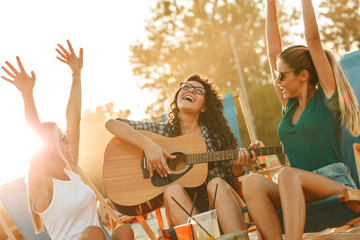 Wall Mural - Group of young female friends sitting on beach on sunbeds,singing and playing guitar.