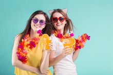 Two Pretty Smiling Caucasian Girls Wears T-shirts With Lei And Sunglasses Hold Orange Cocktails On Blue Background
