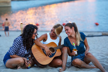 Wall Mural - Group of young female friends sitting on the river beach ,singing and playing guitar.Joying in summer evening .